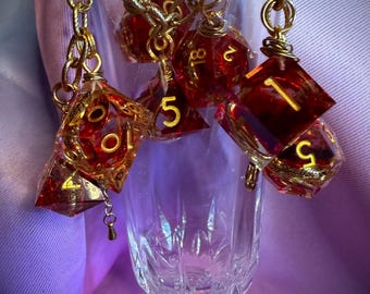 May include: A collection of translucent red polyhedral dice with gold-colored numbers, attached to a gold-toned metal chain draped over the rim of a clear glass flute. The dice feature various geometric shapes including d20 and d10 designs.