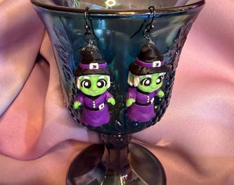 May include: A pair of dangle earrings featuring miniature green-skinned figures wearing purple dresses and black pointed hats with white bands. The figures have white hair and black metal earring hooks attached to the top of the hats.