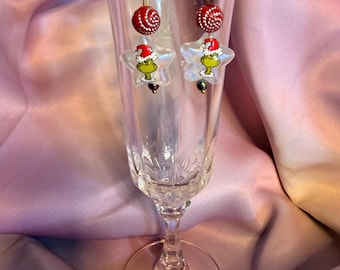 May include: A pair of dangle earrings featuring a green character wearing a red Santa hat inside a clear star-shaped charm. Above the star is a round red bead with white swirls, suspended from a gold-toned metal hook wire.