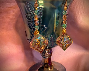 May include: A pair of dangle earrings featuring clear, multi-faceted polyhedral dice with gold-colored numbers. Each earring is suspended from a gold-toned wire hook and accented with a vertical arrangement of small, iridescent teal and amber beads.