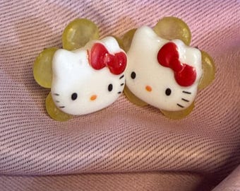 May include: A pair of stud earrings featuring the white Hello Kitty character face with a red bow, set against a translucent yellow flower-shaped backing. The earrings have black eyes, a yellow nose, and black whiskers on each side of the face.