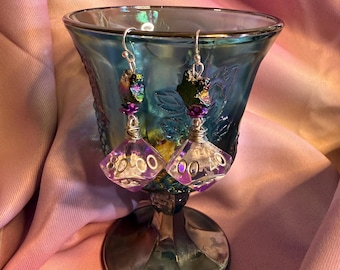 May include: A pair of dangle earrings featuring translucent purple polyhedral dice with white numbers. Each earring is suspended from a silver-toned hook, accented with a small purple flower bead and a dark, iridescent, textured stone-like topper.