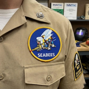May include: Tan military-style shirt with a round patch featuring a cartoon bee holding a weapon, with the word "SEABEES" below. A smaller patch on the sleeve reads "NCF SEABEE RATING". A silver-colored pin is on the collar.