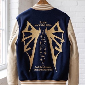 Könnte beinhalten: Eine marineblaue und beige Collegejacke mit goldenen Akzenten. Auf der Rückseite befindet sich ein goldenes Flügeldesign mit dem Text "To the stars who listen" und "And the dreams that are answered". Die Jacke hängt an einem Holzbügel.