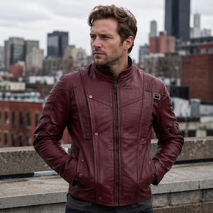 Può includere: Giacca in pelle bordeaux con collo alto e cerniera frontale. La giacca presenta un design trapuntato sulle spalle e sulle braccia, con alcune piccole tasche. La persona è in piedi su un tetto con lo skyline della città sullo sfondo.
