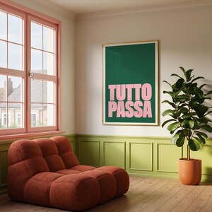 May include: A framed wall art print featuring the text TUTTO PASSA in bold light pink letters against a solid dark green background, displayed on a wall above a low, tufted, rust-colored modular sofa next to a potted green plant.
