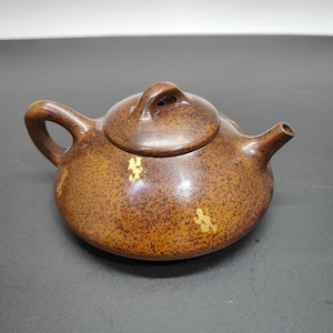 May include: A small, brown teapot with a speckled pattern and a lid. The teapot has a curved handle and a spout. Gold accents are visible on the body. The teapot is made of a dark wood-like material.