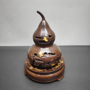 Peut inclure: Un brûle-encens en métal marron en forme de gourde, doté de découpes complexes et d'accents dorés. L'objet repose sur une base circulaire assortie de couleur marron, ornée de motifs dorés sur tout son bord extérieur.