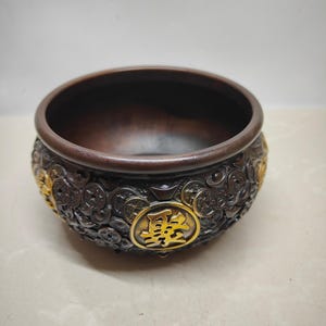 May include: Bronze-colored bowl with intricate coin and character detailing. The bowl features a dark interior and gold-colored accents. The exterior is adorned with embossed coin patterns and a central gold emblem with Chinese characters.