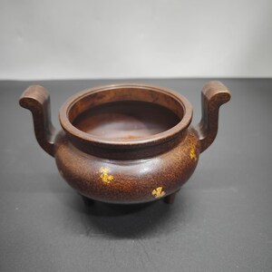 May include: A round, dark brown metal incense burner with a smooth, mottled texture and small, irregular gold-colored flecks scattered across the exterior. It features two curved, upright handles on opposite sides and stands on three short legs.