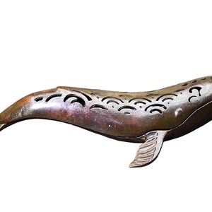 May include: A bronze-toned metal whale figurine featuring a series of cut-out wave patterns along its dorsal side. The sculpture has a smooth, metallic finish with visible texture on the tail and pectoral fin, capturing a swimming motion.