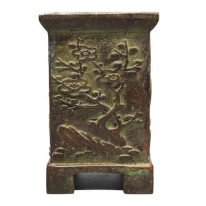 May include: A rectangular bronze-toned planter with a weathered, dark patina. The exterior features a raised relief design of a flowering plum tree with branches, blossoms, and buds. The vessel has a wide rim and a base with two feet.