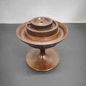 May include: A dark brown, circular wooden incense burner featuring a tiered, pedestal-style base and a wide, shallow bowl top. The center of the bowl contains a raised, concentric circular platform with a small hole for holding incense sticks.