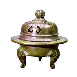 May include: A bronze-toned metal incense burner featuring a rounded body supported by three curved legs. The burner includes a wide, flat rim and a removable domed lid with cut-out decorative patterns and a small, textured knob at the very top.