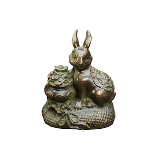 Puede incluir: Una figura de metal en tono bronce que representa un conejo sentado junto a un pequeño recipiente ornamentado con tapa. El conejo presenta patrones de pelaje texturizados en el lomo, mientras que el recipiente descansa sobre una base tejida.