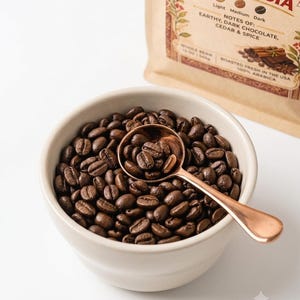 May include: A beige ceramic bowl filled with dark roasted whole coffee beans and a copper-toned metal spoon. Behind the bowl is a tan coffee bag labeled Indonesia, noting flavor profiles of earthy, dark chocolate, cedar, and spice.