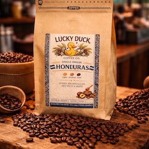 Puede incluir: Una bolsa de papel marrón de café en grano entero Honduras de Lucky Duck Coffee Co., 340g. La etiqueta presenta un pato amarillo con una taza de café y texto que indica miel, azúcar moreno, frutos rojos y sabores a nuez. Los granos de café están esparcidos alrededor de la bolsa.
