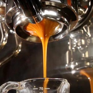 May include: A chrome espresso machine portafilter dispensing a steady stream of dark brown espresso into a clear glass measuring cup with white markings, including a visible 70 ml indicator, while steam rises from the brewing process.
