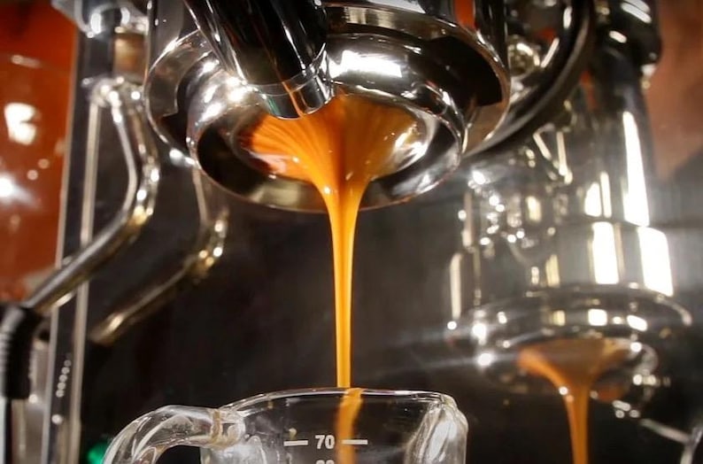 May include: A chrome espresso machine portafilter dispensing a steady stream of dark brown espresso into a clear glass measuring cup with white markings, including a visible 70 ml indicator, while steam rises from the brewing process.