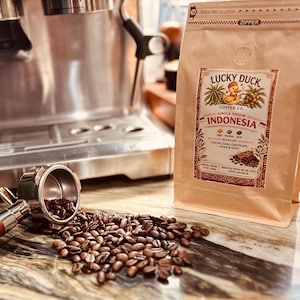 May include: A tan coffee bag labeled Lucky Duck Coffee Co. Indonesia with a duck illustration, next to a metal portafilter spilling roasted coffee beans. The label lists notes of earthy, dark chocolate, cedar, and spice, with a 12 oz weight.