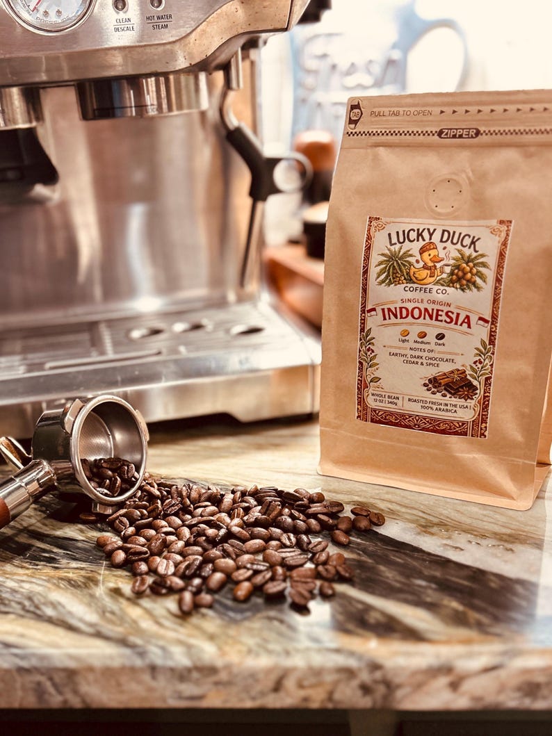 May include: A tan coffee bag labeled Lucky Duck Coffee Co. Indonesia with a duck illustration, next to a metal portafilter spilling roasted coffee beans. The label lists notes of earthy, dark chocolate, cedar, and spice, with a 12 oz weight.