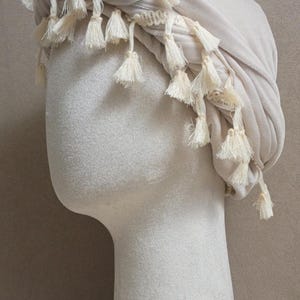 May include: A beige turban-style head covering with cream-colored tassels, displayed on a mannequin head. The fabric appears soft and draped, with the tassels adding a decorative element. The background is a neutral color.