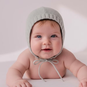 May include: A baby wearing a pale green knitted bonnet with a tie fastening. The bonnet covers the baby's head and ears, with the ties hanging down. The baby has blue eyes and is smiling.