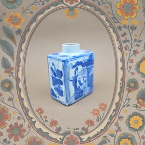 Könnte beinhalten: Eine blau-weiße Porzellan-Teedose mit rechteckiger Form und kleiner zylindrischer Öffnung. Die Dose zeigt detaillierte blaue Illustrationen von Figuren und Blättern. Der Hintergrund ist ein Blumendesign.