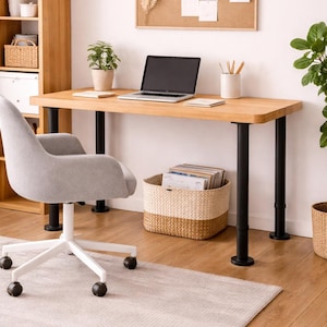 May include: A light wood desk with black legs, a laptop, and a small potted plant. A gray office chair sits on a light gray rug. A woven basket holds magazines. A corkboard with notes and a wooden bookshelf are in the background.