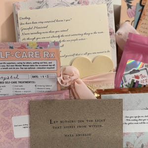 May include: A gift box filled with self-care items. The box contains cards with inspirational quotes, a pink pouch with stickers, and other stationery. The color palette includes pink, beige, and floral patterns, creating a soothing aesthetic.