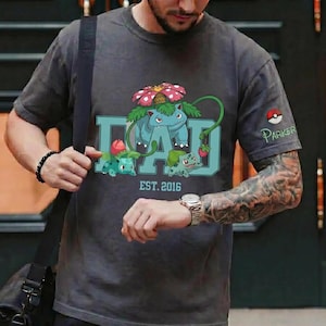 May include: Dark gray t-shirt featuring a blue and green graphic of a cartoon character with the word "DAD" in teal. The shirt also has the text "EST. 2016" and a small logo with the name "Parker".