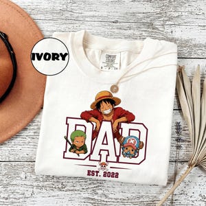 May include: Ivory-colored t-shirt with a graphic of a smiling cartoon character wearing a straw hat, red shirt, and the word "DAD" in maroon. Below the word "DAD" are two more cartoon characters and the text "EST. 2022".