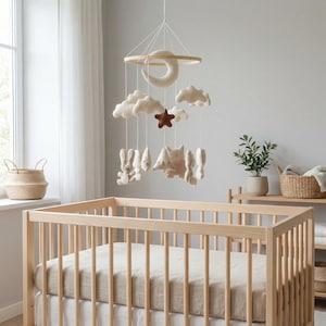 Puede incluir: Una cuna con estructura de madera y colchón beige. Sobre la cuna cuelga un móvil con un aro de madera, una luna creciente, nubes, una estrella y figuras de animales de peluche en colores neutros. La habitación tiene una ventana y una planta.