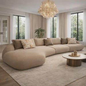 Op de afbeelding: Een grote, beige hoekbank met verschillende kussens in een woonkamer. De bank is gebogen en staat op een lichtgekleurd tapijt. Een ronde salontafel en een kroonluchter zijn ook zichtbaar.