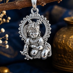 May include: A polished silver pendant featuring a detailed depiction of a deity in a kneeling posture, adorned with traditional jewelry and a headpiece, framed by a circular halo with radiating points, suspended from a simple silver bail.