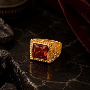 Pode incluir: Um anel de ouro com uma pedra vermelha quadrada. O anel tem uma banda texturizada com um padrão repetitivo. A pedra é definida em uma moldura quadrada com pequenos detalhes decorativos. O anel é exibido em uma superfície escura e texturizada.