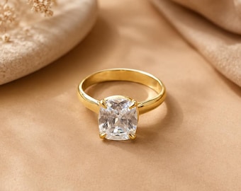 Può includere: Un anello in oro con una singola grande pietra preziosa trasparente a taglio cuscino, fissata in una montatura a quattro griffe. La fascia è liscia e lucida, caratterizzata da un design classico con una pietra sfaccettata e prominente.