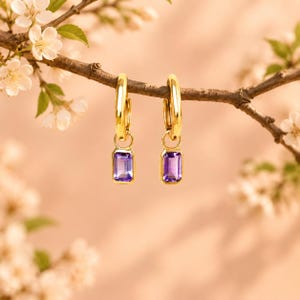 Könnte beinhalten: Ein Paar goldene Creolen, die jeweils mit einem rechteckigen, violetten Amethyst-Edelsteinanhänger versehen sind, der an der Unterseite hängt. Die Ohrringe sind poliert und haben eine glatte, reflektierende Oberfläche.