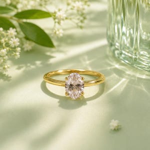 Peut inclure: Une bague en or ornée d'une pierre précieuse transparente taille ovale, maintenue par un sertissage à quatre griffes. L'anneau est lisse et poli, présentant un design classique avec une pierre centrale qui reflète la lumière.