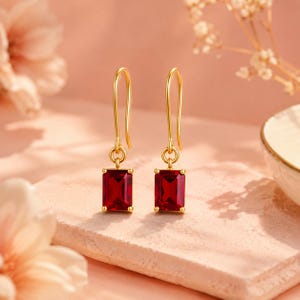 Peut inclure: Une paire de boucles d'oreilles pendantes en or avec des pierres précieuses rectangulaires à facettes rouge foncé. Chaque boucle est suspendue à un crochet en or simple, mettant en valeur une finition polie sur le métal et une teinte rouge vive.