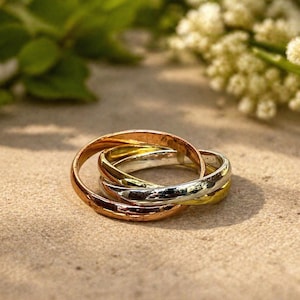 Könnte beinhalten: Drei ineinandergreifende Ringe in Gold, Silber und Roségold. Die Ringe sind ineinander verschlungen und bilden ein einzigartiges Design. Die Ringe sind vor einem neutralen Hintergrund mit verschwommenem Grün und weißen Blumen im Hintergrund platziert.