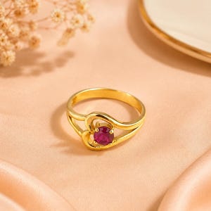 Puede incluir: Un anillo de oro con un engaste en forma de corazón que sostiene una piedra preciosa redonda de color rosa intenso. La banda es lisa y pulida, con el diseño de corazón integrado en la parte superior, creando una estructura metálica fluida.