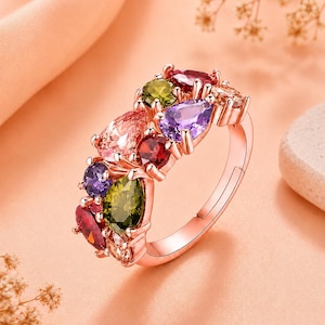 Op de afbeelding: Een roségouden ring met een cluster van peervormige edelstenen in diverse kleuren, waaronder rood, paars, olijfgroen en lichtroze. De stenen zijn gezet in een klauwzetting langs de bovenste helft van de ringband, die een gladde afwerking heeft.