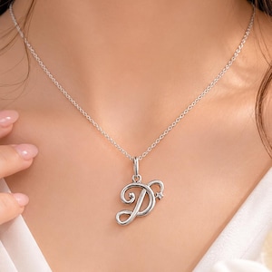 Op de afbeelding: Een zilveren ketting met een hanger in de vorm van de letter "D". De hanger heeft een cursief ontwerp met een kleine ster. De ketting wordt om de nek gedragen.