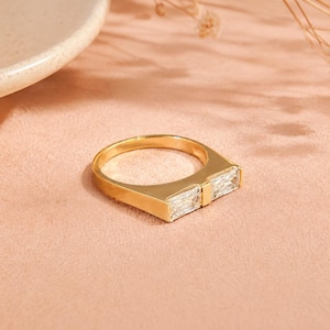 Op de afbeelding: Een goudkleurige ring met een plat, breed bandontwerp en twee rechthoekige, heldere stenen in baguette-slijpvorm die horizontaal naast elkaar in het midden zijn geplaatst. Het metaal heeft een gladde, gepolijste afwerking en de stenen zijn gefacetteerd.