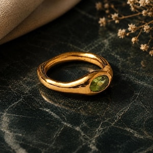 Op de afbeelding: Een gouden ring met een gladde, getextureerde band en een groene, ovaalvormige edelsteen. De ring is geplaatst op een donkere, getextureerde ondergrond, met een zachte, neutrale stof op de achtergrond.
