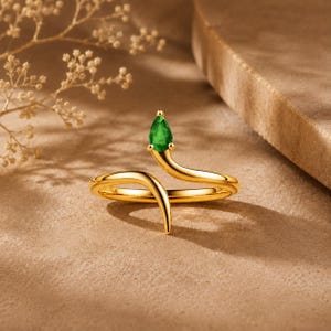 May include: A gold-toned open ring featuring a smooth, curved band that tapers at both ends. One end of the band is topped with a single, pear-shaped, vibrant green gemstone set in a small, four-prong gold-toned metal setting.