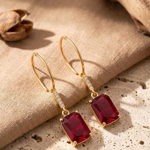 Könnte beinhalten: Ein Paar goldene Creolen mit einem rechteckigen, tiefroten Edelsteinanhänger. Jeder Ohrring verfügt über einen dünnen, goldenen Verbindungssteg, der mit kleinen, klaren, funkelnden Steinen besetzt ist und den Reifen mit dem facettierten Anhänger verbindet.