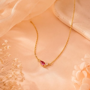 Op de afbeelding: Een delicate gouden ketting met een kleine hanger met een rode edelsteen in marquise-slijpvorm en een enkele ronde, heldere steen. De ketting is gemaakt van een dunne, gepolijste goudkleurige metalen ketting die aan de centrale zetting vastzit.