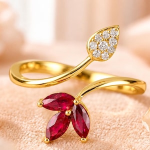 May include: A gold bypass ring featuring a leaf-shaped accent set with small clear round stones and a three-petal flower motif set with marquise-cut deep red stones. The band is polished gold, creating a delicate and open design for the finger.